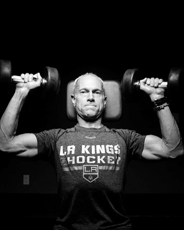 Rick Scott pressing two dumbbells overhead in an LA Kings Hockey shirt